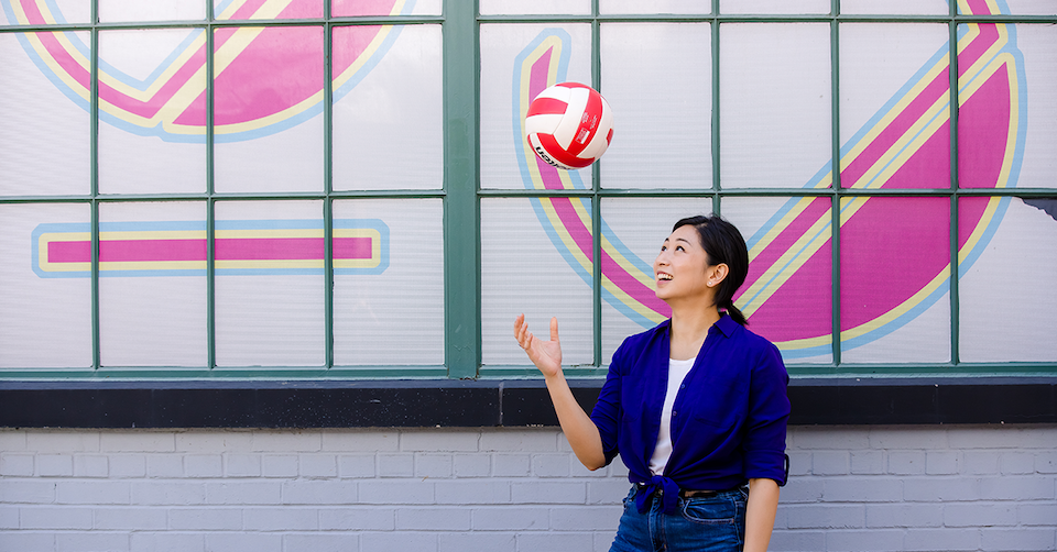 Capital One Tech associate Jessica stands in front of a grey and pink wall outside and throws a volleyball in the air